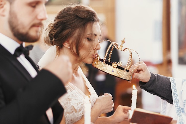 Braut und Bräutigam küssen goldene Kronen aus Priesterhand während der Hochzeitszeremonie Spirituelles Paar Hochzeitsehe in der Kirche Emotionale romantische Momente