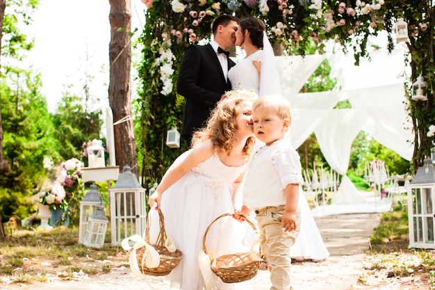 Foto braut und bräutigam küssen sich nach der hochzeitszeremonie. entzückende kinder mit fliegenden rosenblättern in körben