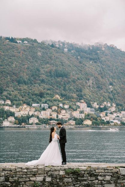 Braut und Bräutigam stehen Hand in Hand und schauen einander an auf einem Steinpier in der Nähe des Lake Como in Italien