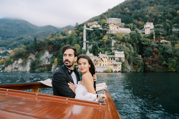Braut und Bräutigam umarmen sich, während sie vorwärts schauen, während sie auf einer Yacht stehen, die auf dem Como-See segelt.