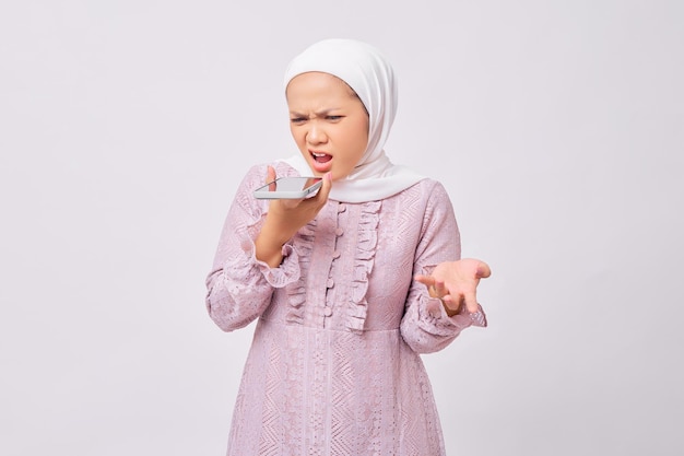 Brava e bonita jovem muçulmana asiática usando hijab e vestido roxo gritando alto no celular isolado no fundo branco do estúdio