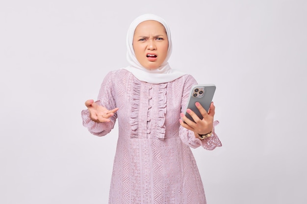 Brava linda jovem muçulmana asiática usando hijab e vestido roxo usando smartphone gritando alto ao receber más notícias isoladas no fundo branco do estúdio