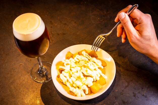 Bravas mit Aioli-Sauce mit einem Bier auf schwarzem Hintergrund, auf einem blauen Teller