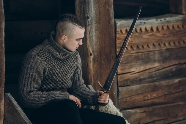 Bravo guerreiro segurando a espada perto do edifício histórico do castelo de madeira na floresta de inverno na escandinávia, bonito viking com moicano posando com arma antes do conceito de cosplay de fantasia de batalha