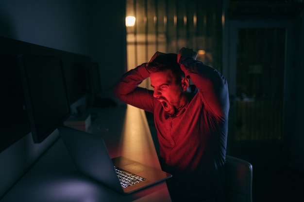 Bravo jovem caucasiano barbudo empregado olhando para laptop enquanto está sentado no escritório tarde da noite.