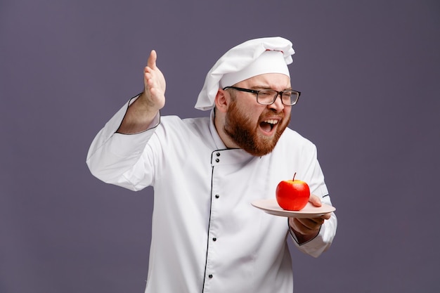 Bravo jovem chef vestindo óculos uniforme e boné segurando a maçã no prato olhando para o lado levantando a mão gritando isolado no fundo roxo