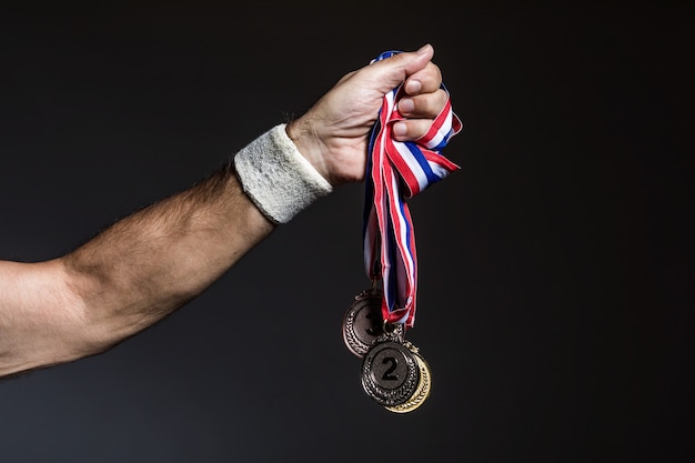 Brazo de atleta anciano sosteniendo tres medallas de oro, plata y bronce sobre un fondo oscuro. Concepto de deportes y victoria.