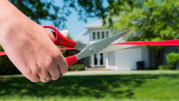Un brazo cortado con tijeras corta una cinta roja y una típica casa estadounidense