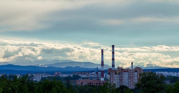 Breites Panorama der modernen städtischen Baustelle des Wohnwohnungsviertels in der Stadt mit grünen Bäumen, Arbeitsturmkränen, hohen Rohrschornsteinen unter blauem Himmel auf entferntem Gebirgszug