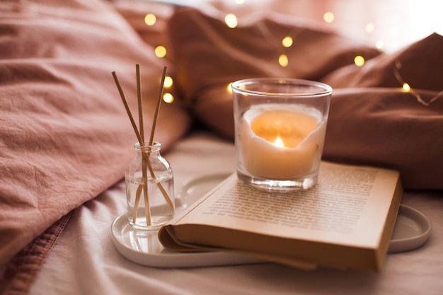 Brennende Kerze mit Aromasticks in Flasche auf Tablett mit offenem Buch im Bett über glühender Weihnachtsbeleuchtung hautnah. Gemütliche Atmosphäre zu Hause. Guten Morgen. Selektiver Fokus.