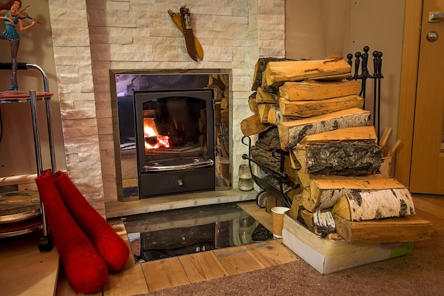 Brennholz in der Nähe von brennendem Kamin. Stiefelständer aus rotem Filz. Zubehör steht Marmor in der Nähe der Hauswand.