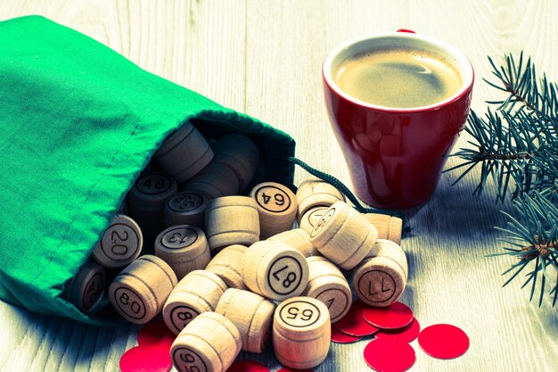 Brettspiel Lotto. Hölzerne Lottofässer mit grüner Tasche, Spielkarten, roten Chips und Tasse Kaffee, Weihnachtstannenzweigen. Farbtoneffekt.