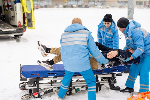 Brigade von Sanitätern in Winteruniform, die bewusstlosen Mann tragen und auf Trage legen, um ihn zum Krankenwagen zu bringen