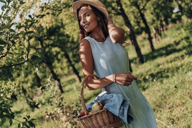 Brillando con belleza natural. Mujer joven atractiva que lleva una canasta con manzanas y apartar la mirada con una sonrisa mientras está de pie en el jardín