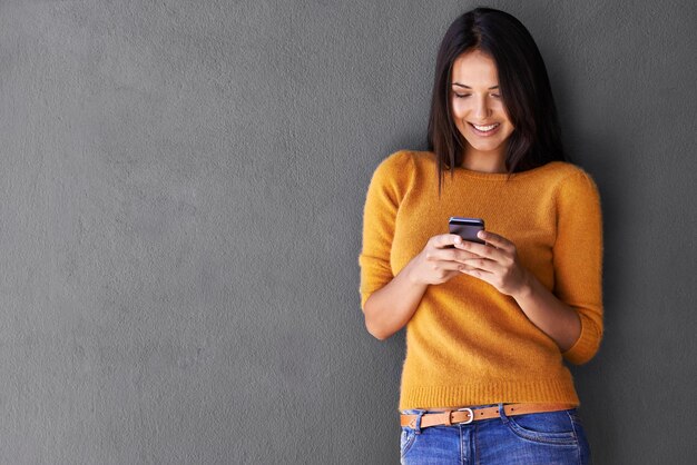 Brillante y burbujeante con buenas noticias Una mujer joven atractiva que usa un teléfono móvil mientras está de pie contra una pared gris