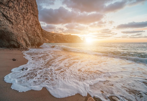 Brillante puesta de sol en el mar las olas chocan contra la roca iluminada por la cálida puesta de sol arena y guijarros volcánicos