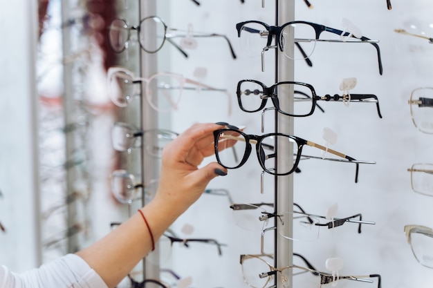 Brille wählen. Kunde in der Optik. Selektiver Fokus zur Hand. Frau, die Korrekturbrille auf Ständer mit Brille berührt. Breites Sortiment im Shop.