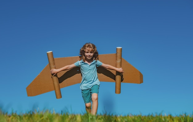 Brincadeira feliz com asas de papelão de avião de brinquedo contra garoto de céu azul se divertindo no campo de verão