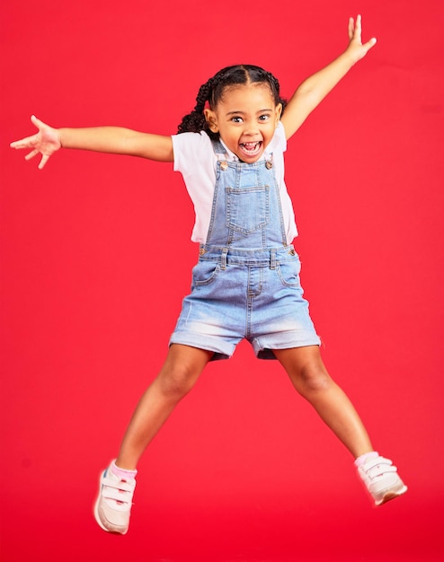 Brincalhão animado e retrato de uma menina pulando enquanto isolada em um fundo vermelho em um estúdio Sorriso jovem e criança com liberdade, energia e felicidade enquanto brincava com um salto em um pano de fundo