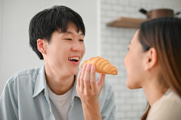 Brincalhão namorado asiático alimentando um croissant para sua namorada na cozinha