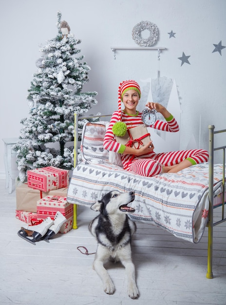 Brincalhão sorridente feliz menino bonito vestido de pijama listrado com presente, relógio e cachorro sentado no quarto decorado de ano novo em casa. Bom humor de Natal. Conceito de estilo de vida, família e férias 2018