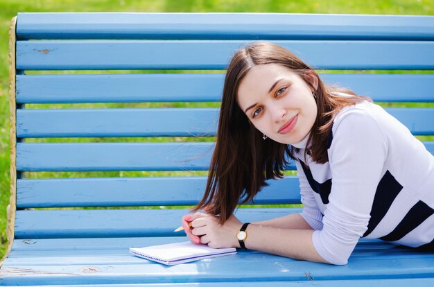 Brinette linda garota sentada num banco, sonhando e escrevendo seus planos em um notebook, relaxando em um parque em um dia ensolarado de primavera