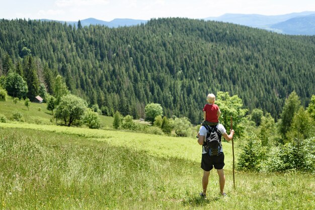 Bringen Sie mit dem Sohn auf seinen Schultern hervor, die mit Personal im grünen Wald, in den Bergen und im Himmel mit Wolken stehen. Rückansicht