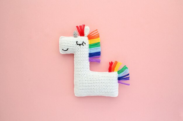 Brinquedo de unicórnio branco macio recheado de crochê amigurumi feito à mão com crina de arco-íris em fundo rosa. Mão