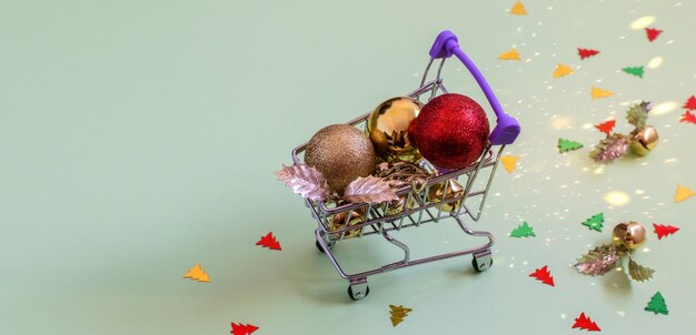 Foto brinquedos de natal brilhantes. bolas douradas e vermelhas, bugigangas e confetes no mini carrinho de compras.