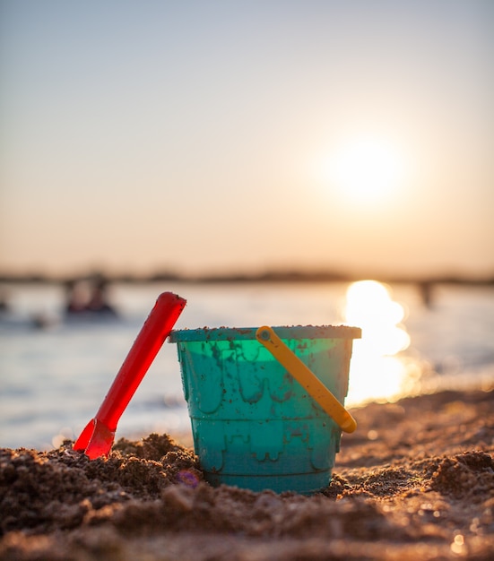 Brinquedos infantis para brincar na areia. Balde de plástico e ancinho na praia ao pôr do sol. O conceito de verão, férias em família e férias.