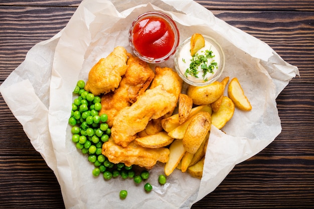 Britischer nationaler traditioneller Fast-Food-Fisch und Chips mit verschiedenen Dips, frischen Erbsen, auf Papier, rustikalem braunem hölzernem Hintergrund