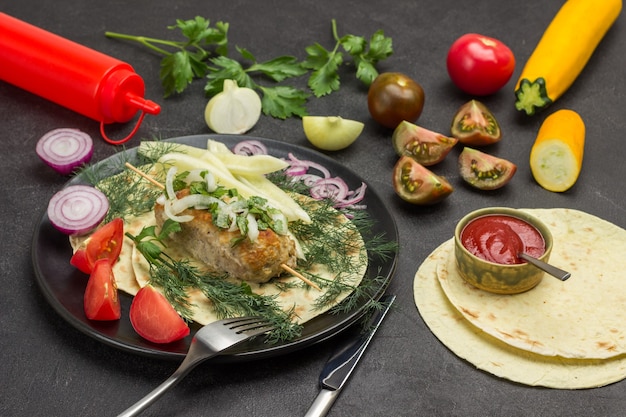 Brocheta de carne con verduras y hierbas en un plato. Pita y tomates picados en la mesa. Fondo negro. Vista superior