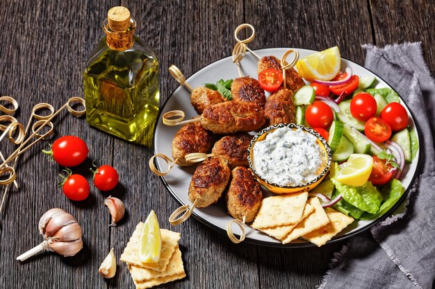 Brochetas picantes de cordero y feta kofta con tomates frescos, menta, rodajas de cebolla roja, galletas saladas y salsa tzatziki en una mesa de madera oscura, vista superior, primer plano