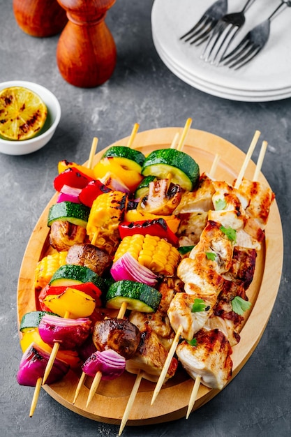 Brochetas de verduras y pollo a la parrilla con maíz dulce pimentón calabacín cebolla tomate y champiñones en un plato de madera