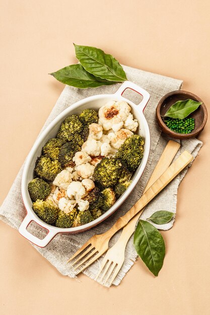 Brócoli y coliflor al horno. Cubiertos de madera, hojas de laurel frescas, pimienta verde. Concepto de comida sana, fondo beige claro, vista superior