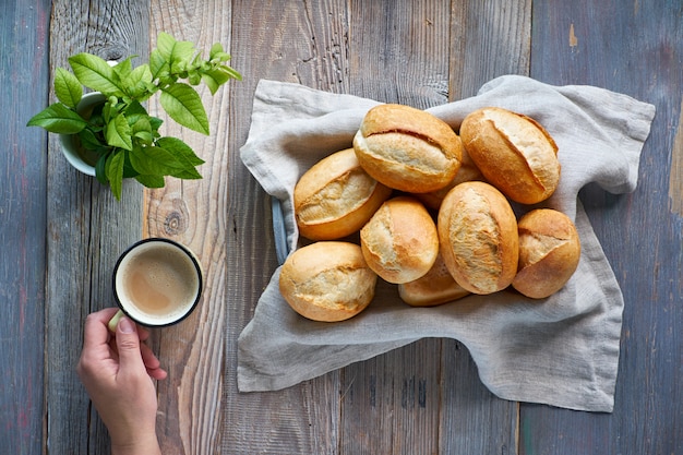 Brötchen im Korb auf rustikalem Holz, Frühlingsblättern und Hand mit Tasse Kaffee