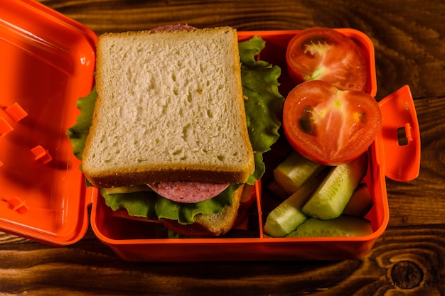 Brotdose mit Sandwich, Gurken und Tomaten auf rustikalem Holztisch