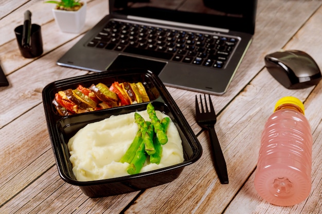 Brotdosenbehälter mit Kartoffelpüree, Spargel und Gemüse auf Holztisch mit Laptop.