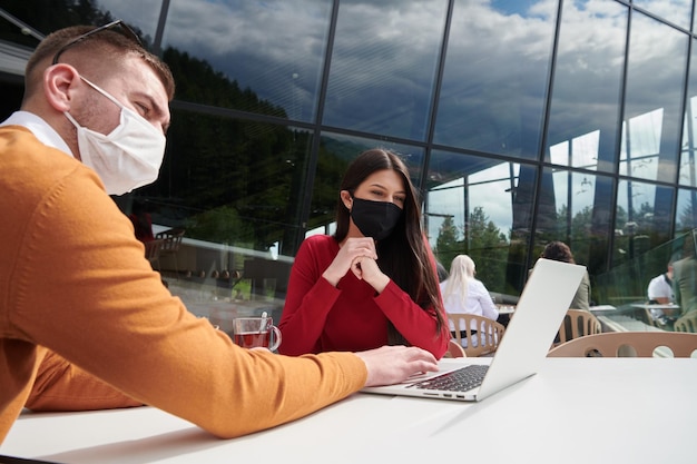 brote de coronavirus Grupo de personas de negocios informales en un restaurante al aire libre con máscara médica protectora, equipo de negocios colaborando e intercambiando ideas de negocios mientras trabajan en una computadora portátil