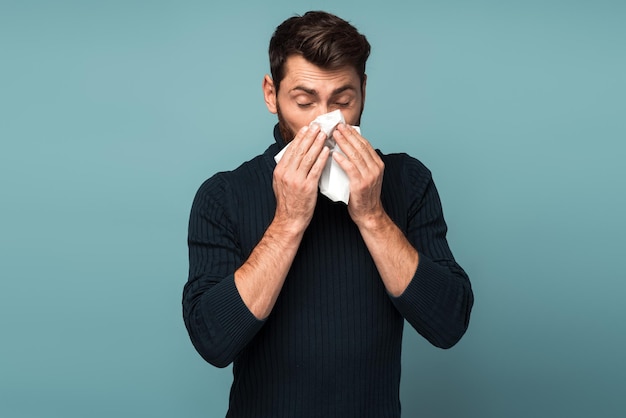 Brote de virus epidemia de gripe Hombre estornudando en la limpieza de la servilleta secreción nasal y tos sufriendo síntomas de gripe fiebre Estudio interior tiro aislado sobre fondo azul