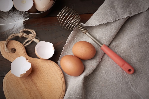 Brown huevos de gallina cruda, cáscaras, tabla de cortar y batidor de mano