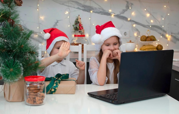 Bruder und Schwester in Weihnachtsmütze haben Spaß. Kinder am Tisch mit Laptop und Geschenken, Freunde oder Verwandte per Videolink anrufen und winken. Familie feiert Weihnachten zu Hause. Soziale Distanzierung.