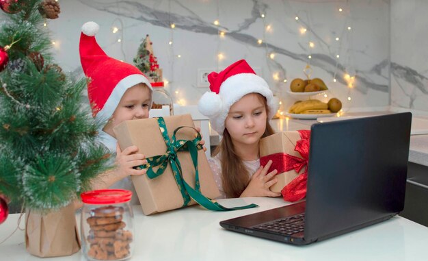 Bruder und Schwester in Weihnachtsmütze haben Spaß. Kinder am Tisch mit Laptop und Geschenken, Freunde oder Verwandte per Videolink anrufen und winken. Familie feiert Weihnachten zu Hause. Soziale Distanzierung.