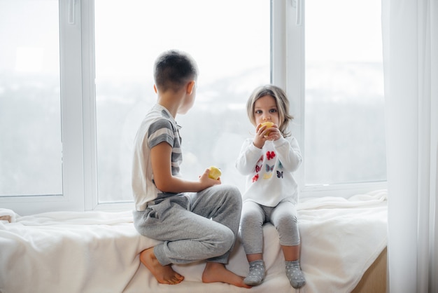 Bruder und Schwester sitzen auf der Fensterbank und spielen und essen Äpfel. Glück.