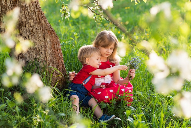 Bruder und Schwester sitzen in einem Park unter einem Baum zwischen den Blumen und lächeln
