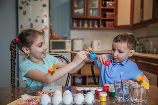 Brünette Brüder und Schwestern schmücken Ostereier, die zu Hause in der Küche am Tisch sitzen