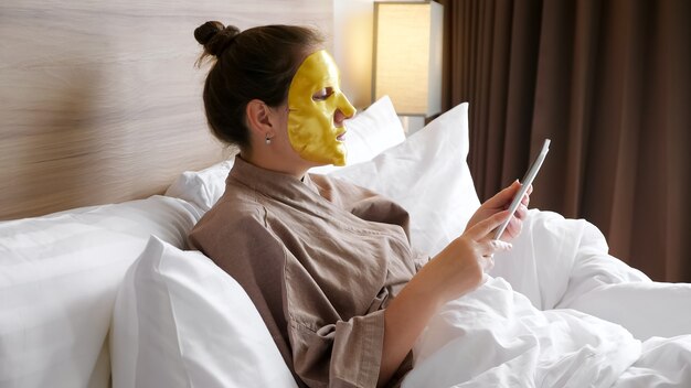 Brünette Dame mit goldener Gesichtsmaske surft im Internet mit Tablet im weichen Bett im komfortablen Hotelzimmer