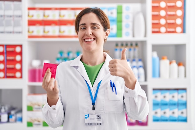 Brünette Frau, die in einer Apotheke arbeitet und ein Kondom hält, das glücklich und positiv lächelt, den Daumen nach oben macht und ein Genehmigungszeichen macht
