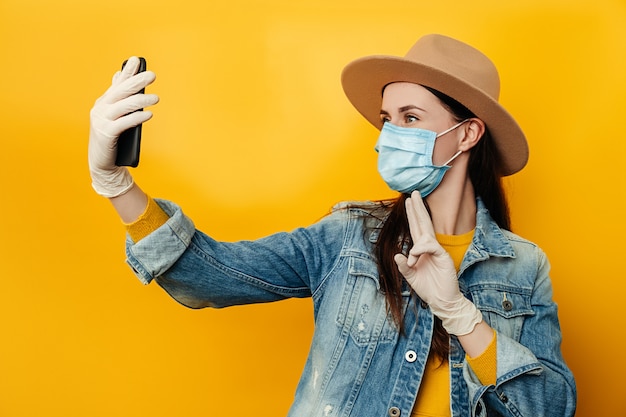 Brünette Frau, die Telefone in Händen hält, macht Selfie, vor gelbem Hintergrund, gekleidet in Jeansjacke, trägt medizinische Schutzmaske, um sich vor Coronavirus zu schützen. Soziales Distanzierungskonzept