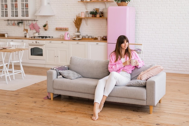 Foto brünette frau genießt einen gemütlichen morgen mit einer tasse tee sitzend auf einem bequemen sofa stilvolle helle küche mit rosafarbenem kühlschrank